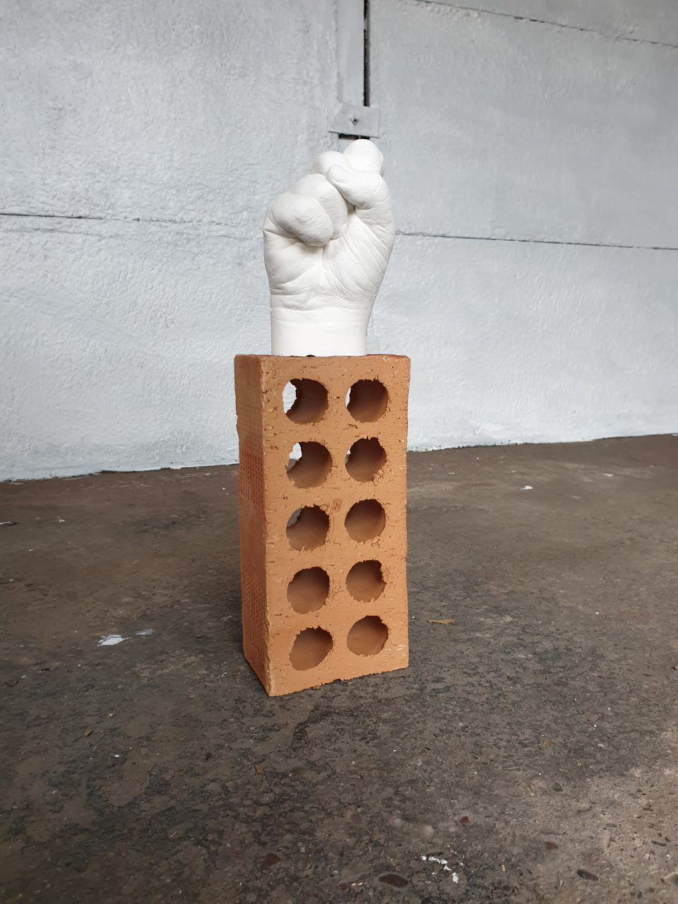 Single white alginate plaster cast of hand in a fist on single orange building brick. 16 x 12 x 8 inches.