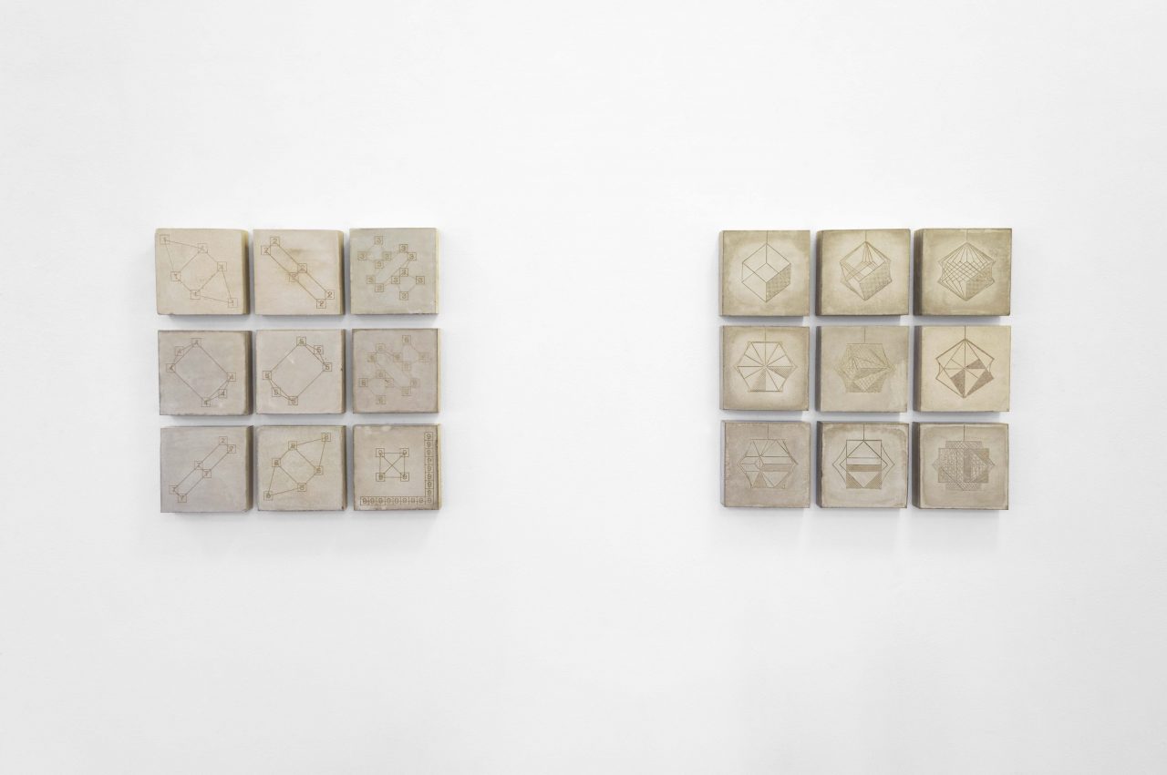 An installation shot of 12 concrete square sculptures hung on a white wall in two sets of 9 square grids. On each of the squares surface is a laser engraved pattern, such as geometric shapes, star patterns and numbers.