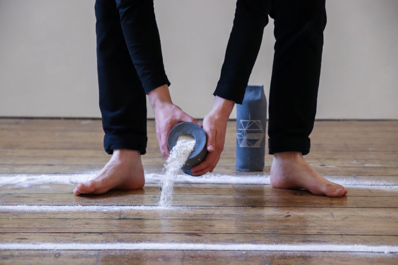 A still image from a performance depicting the artists dressed in black clothes with bare feet. He is hunched over and in his hands he is holding a ceramic pot, in in the process of pouring salt onto the floor forming a salt circle. On the floor behind the performer is another tall ceramic pot glazed grey with a carved geometric design of a spinning square in the centre.