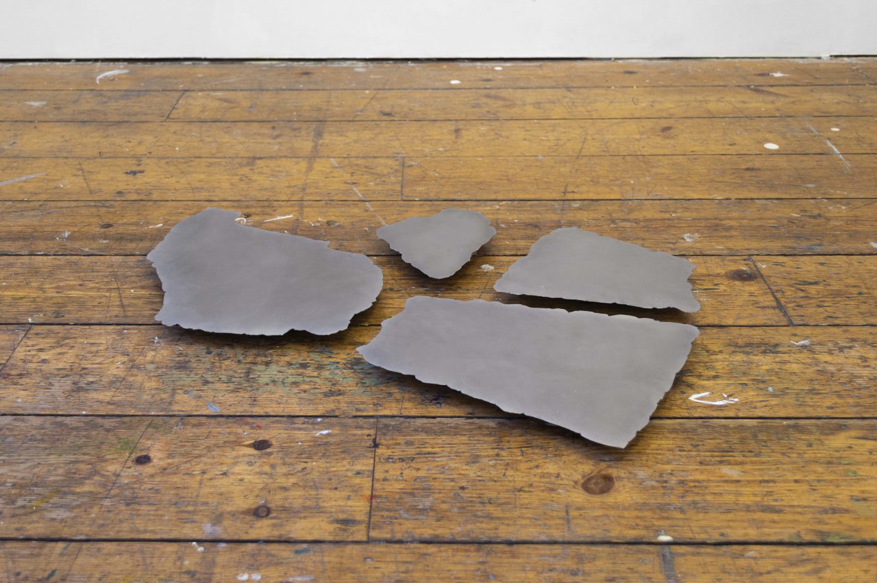 A gallery installation shot of a floor sculpture consisting of 4 polished sheets of steel that has been cut into natural rock lock forms. The sheets appear to float a few cm off of the ground, casting a small shadow below.