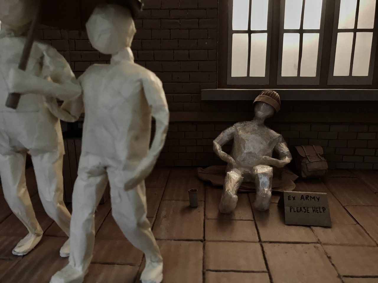 Still taken from stop motion animation of cardboard set and masking tape figures. Two figures walk past another figure who they ignore. Third figure is made from clear tape and sits on the pavement with a leg outstretched.