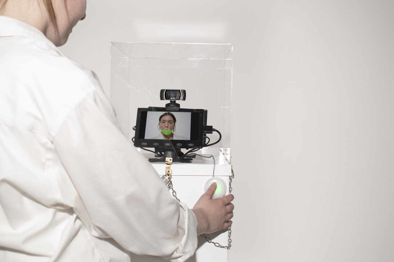 Image taken over the shoulder of a person who is pressing a button on the side of a white plinth. On this plinth sits a screen on which you can see the persons face. The display of their face is partially covered by a green calibration animation.